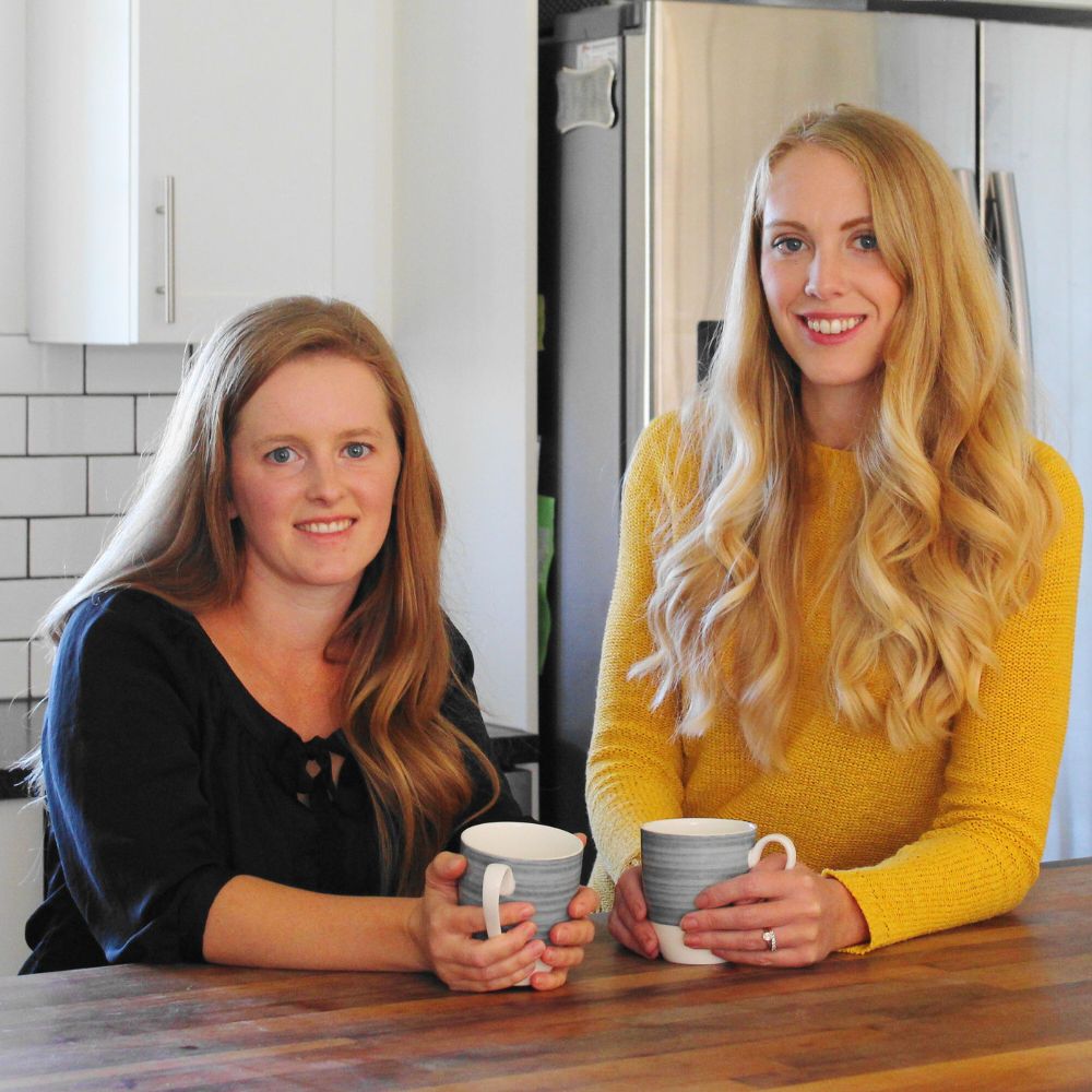 two women standing at a kitchen counter, holding coffee cups and smiling at the camera