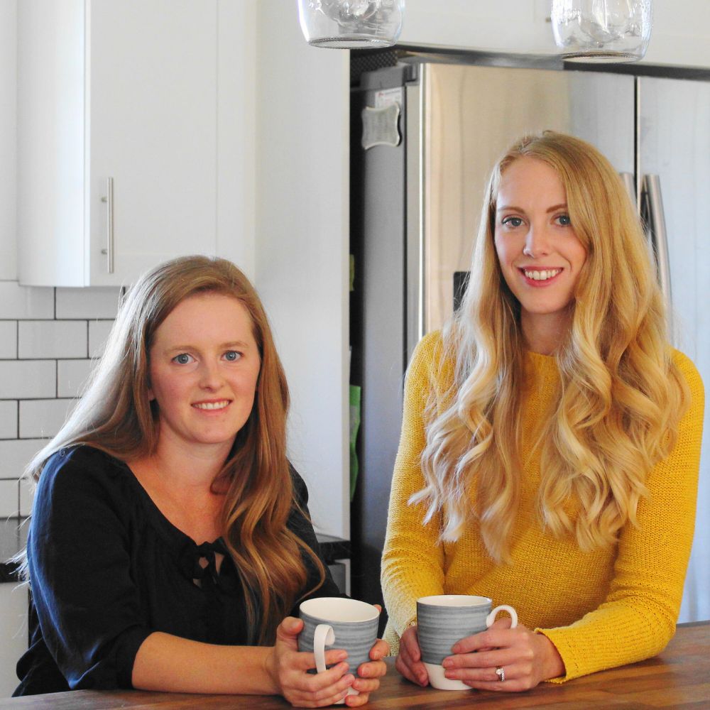 two women standing at a kitchen counter, holding coffee cups and smiling at the camera