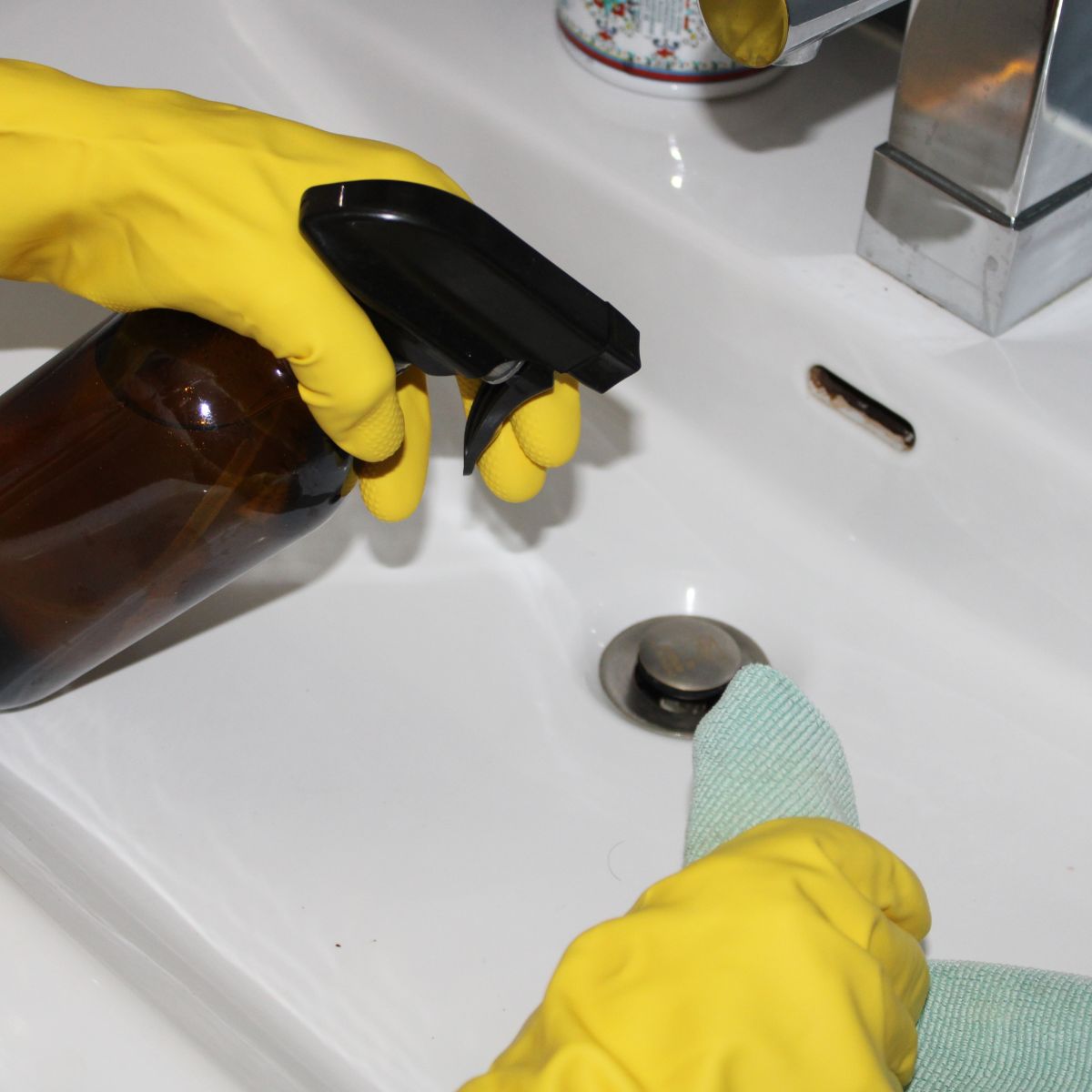 hands wearing cleaning gloves over a sink with a spray bottle in one hand and a cloth in the other