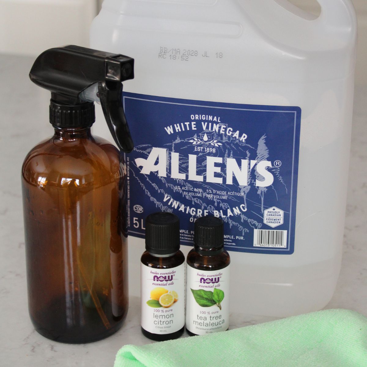 amber glass spray bottle next to a bottle of vinegar, and small essential oil bottles, and a microfiber cloth