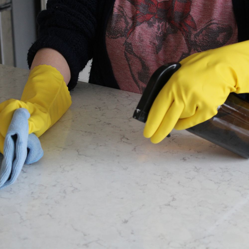 woman wearing cleaning gloves holding a glass spray bottle in one hand and a cloth in the other