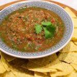 A bowl of fresh homemade salsa garnished with a cilantro leaf, served with tortilla chips.