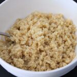 A bowl of cooked white quinoa, showcasing its fluffy texture, served with a spoon in a white bowl against a black background.