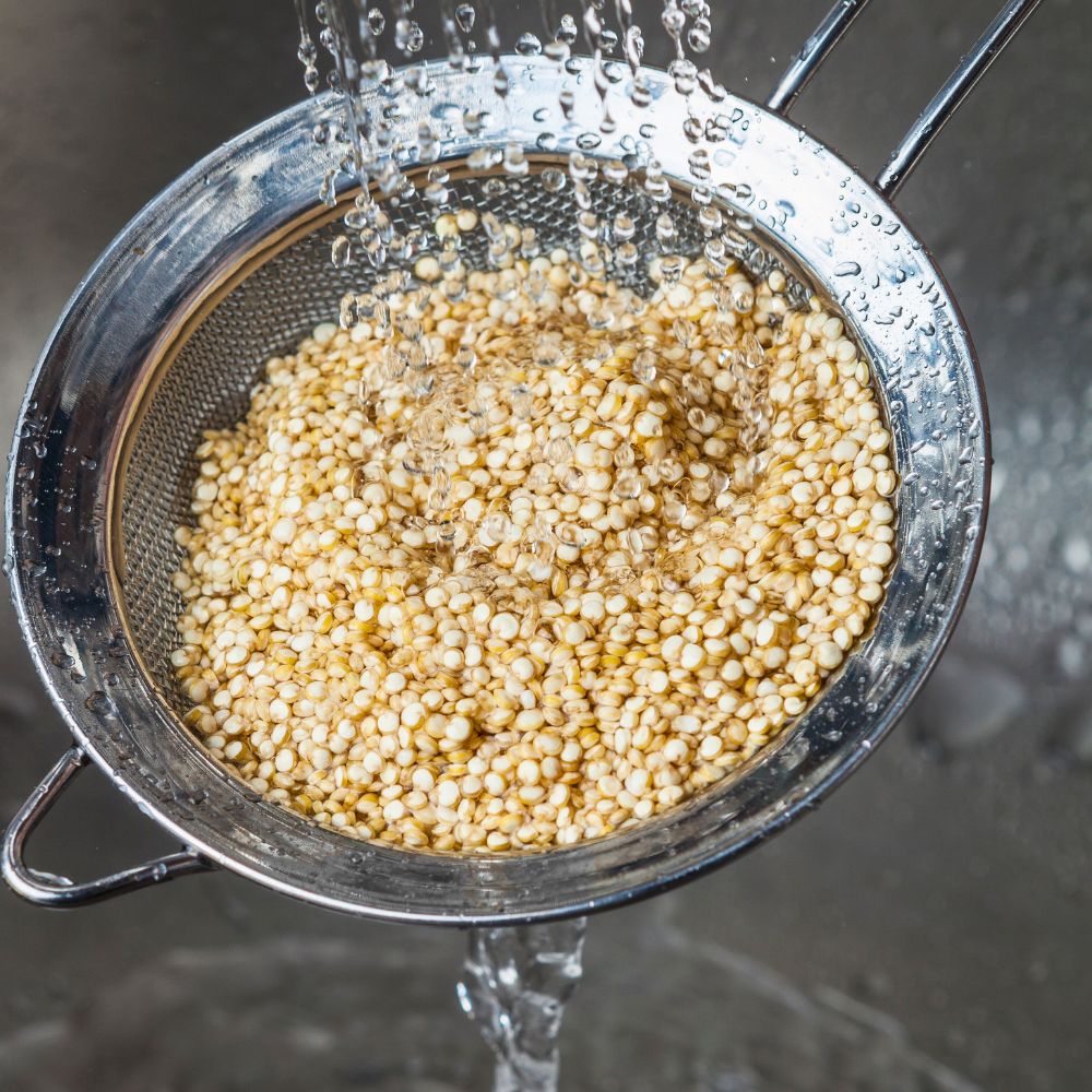 White quinoa being rinsed in a fine mesh strainer under running water over a sink, with water dripping out the bottom to remove bitterness for better taste.