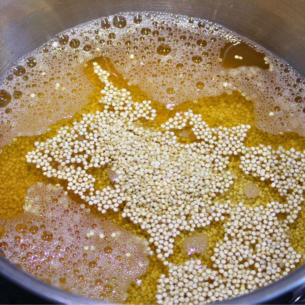 White quinoa and bone broth combined in a medium saucepan on a stove at the beginning of the cooking process, with raw quinoa floating on the surface.