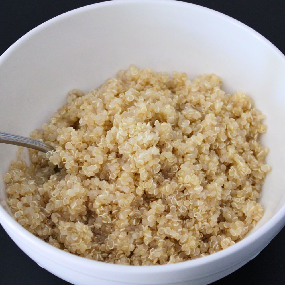 A bowl of cooked white quinoa, showcasing its fluffy texture, served with a spoon in a white bowl against a black background.