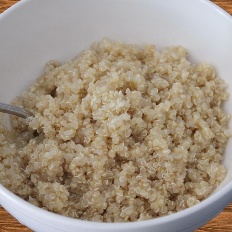 A bowl of cooked white quinoa, showcasing its fluffy texture, served with a spoon in a white bowl against a black background.