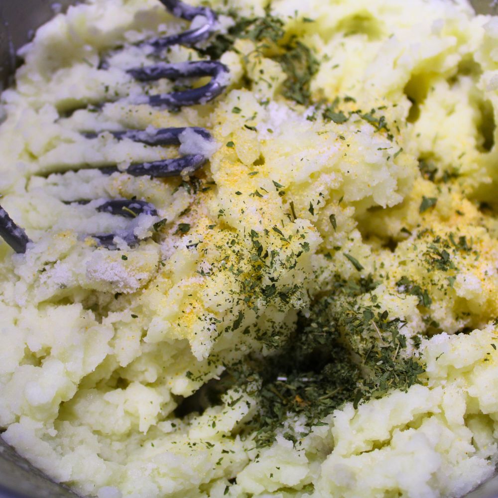 mashed potatoes in a pot with a potato masher, seasoning poured onto potatoes but not yet mixed in