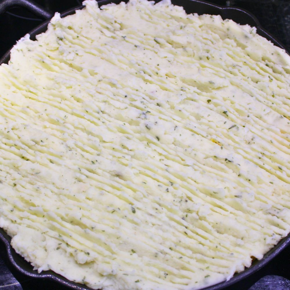 mashed potatoes with fork lines for texture, spread over ground beef in homemade shepherd's pie in a cast iron skillet