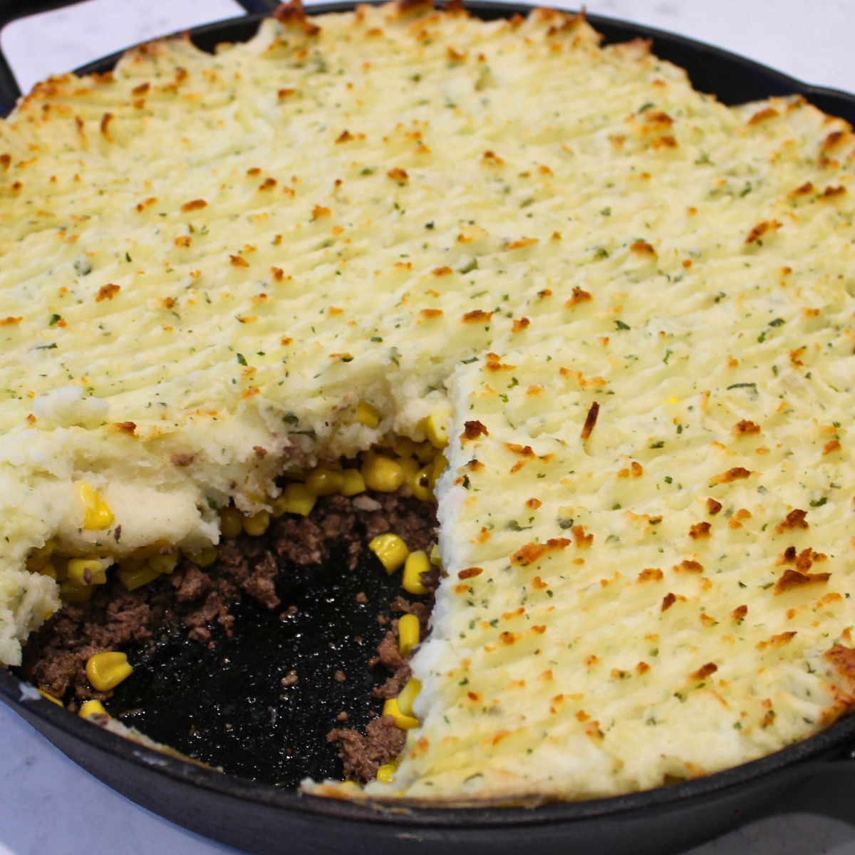 homemade shepherds pie in a cast iron skillet with a slice removed so that the beef and corn filling are visible