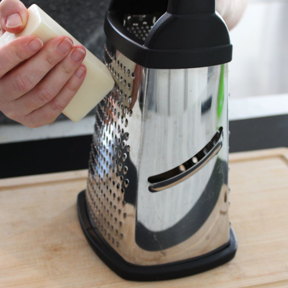 A person grating a bar of laundry soap using a box grater on a wooden cutting board, preparing ingredients for homemade laundry detergent.
