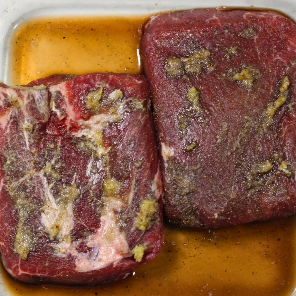 Two raw cuts of steak marinating in olive oil, minced garlic, salt, and pepper inside a white baking dish before oven-baking in a cast iron skillet.