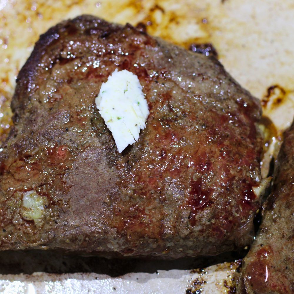 A cooked juicy steak with seared edges, topped with a small dollop of herb butter, resting in an enameled cast iron pan.
